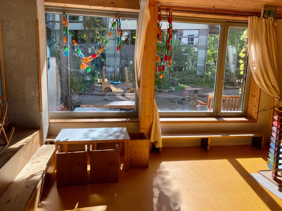 Sonniger Gruppenraum mit großen Fenstern, bunten Papierketten und Blick in den Garten