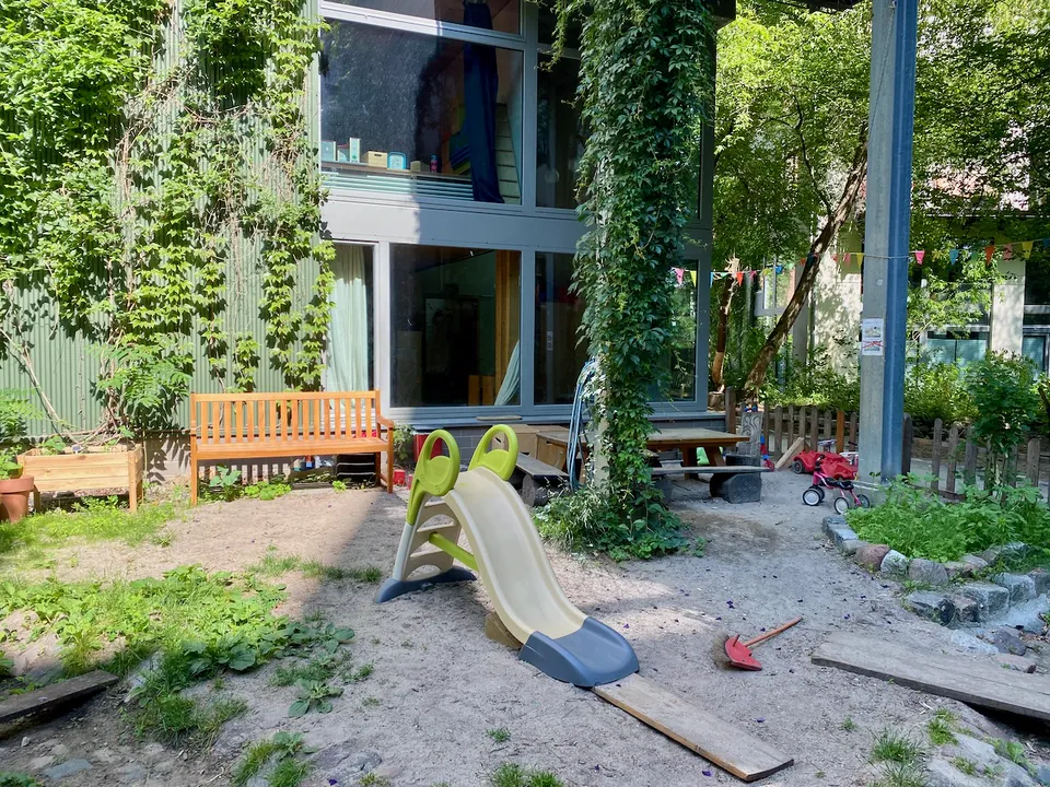 Der Garten des Kinderladens mit Rutsche, Holzbank, Sandfläche und Kletterpflanzen an der Hauswand